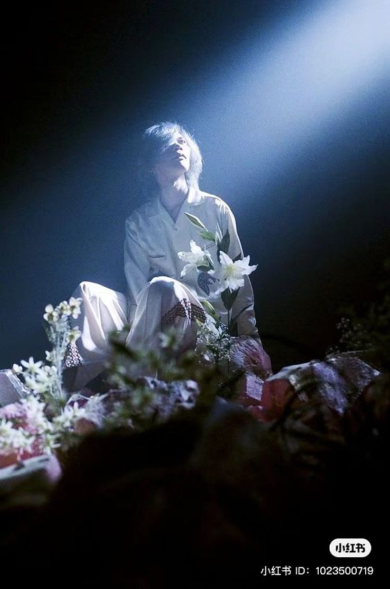 a man in a white silk outfit sits on a mound of flowers and crystals, bathed in a single blue spotlight that shines down from above. the background is dark, emphasizing the dramatic lighting and creating a mystical, dreamy atmosphere.  he is looking up towards the light source, and is holding a white lily in his hand.