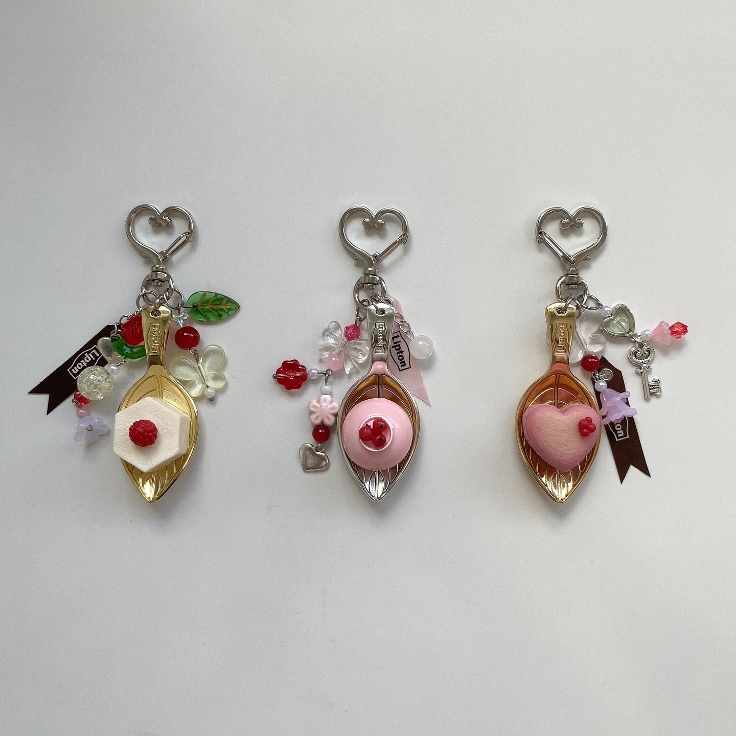 the image shows three handcrafted spoon keychains. each keychain features a small spoon pendant filled with a different clay dessert or treat. each has a silver, heartshaped key ring. they are decorated with different colored beads and charms. each has a small lipton ribbon tied to the ring. the keychains appear to be handcrafted or novelty items.