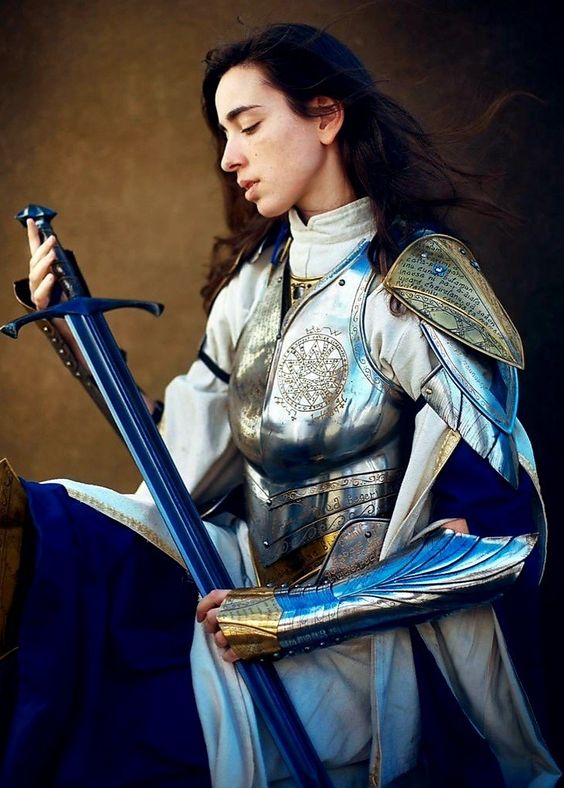 a woman with long dark hair, clad in ornate silver and gold armor, sits in profile holding a sword. her head is bowed slightly, eyes closed, as if in contemplation or prayer. the armor features intricate detailing, including winged pauldrons and a breastplate adorned with a circular design. a blue tunic peeks out from beneath the armor. the background is a warm brown, suggestive of an indoor setting. the image evokes a sense of strength, grace, and inner peace.