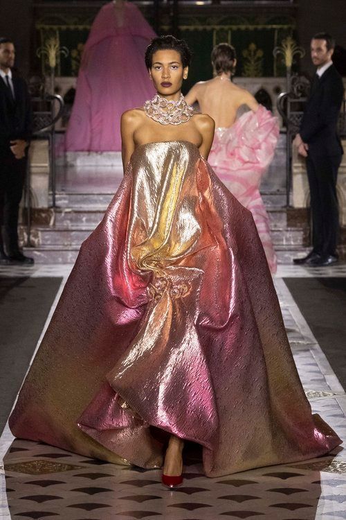 a female model walks the runway wearing a beautiful, shimmering, gold and pink ombre gown. the dress has a unique shape with a dramatic, flowing skirt and an interesting, almost sculpturelike design. she is wearing a chunky statement necklace and simple makeup.  the model is walking towards the camera with a confident expression.  the background is a classic, elegant setting, perhaps a fashion show.  the overall image is glamorous and sophisticated.