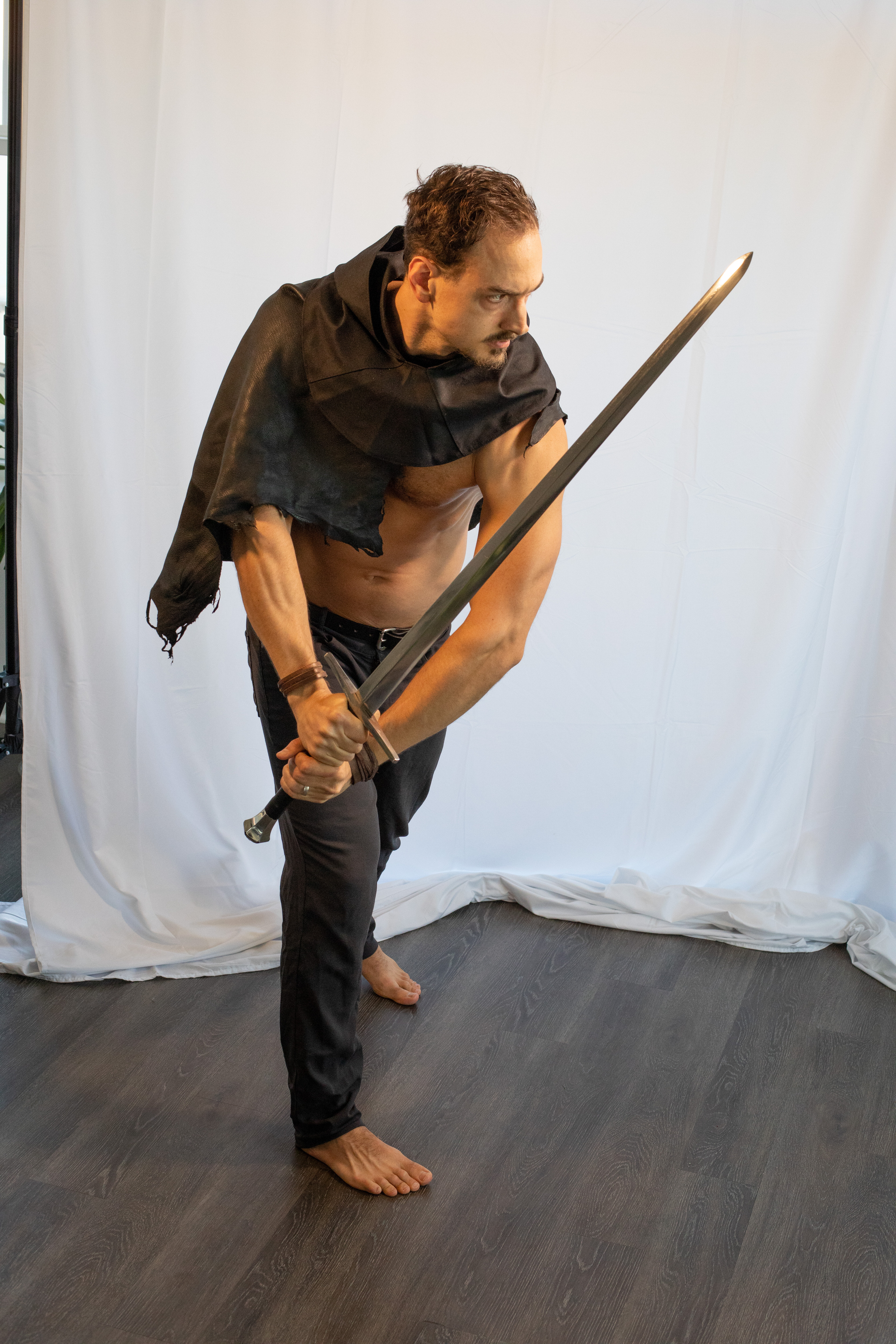 a muscular man with a determined expression stands in a fighting stance. he is shirtless except for a dark, flowing cape draped over his shoulders. he grips a long, gleaming sword with both hands, its point directed towards something offscreen. the setting appears to be a studio, with a plain white backdrop and dark wooden floor. the lighting is bright and artificial, highlighting the man's physique and the shine of the sword.