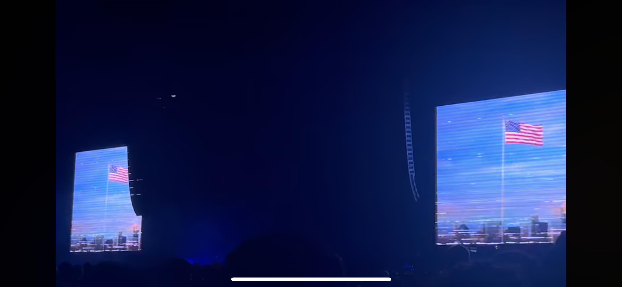 two large screens show an american flag flying in front of a city skyline. the screen is in a dark concert venue with an audience watching.  there is a dark figure standing in front of the left screen.