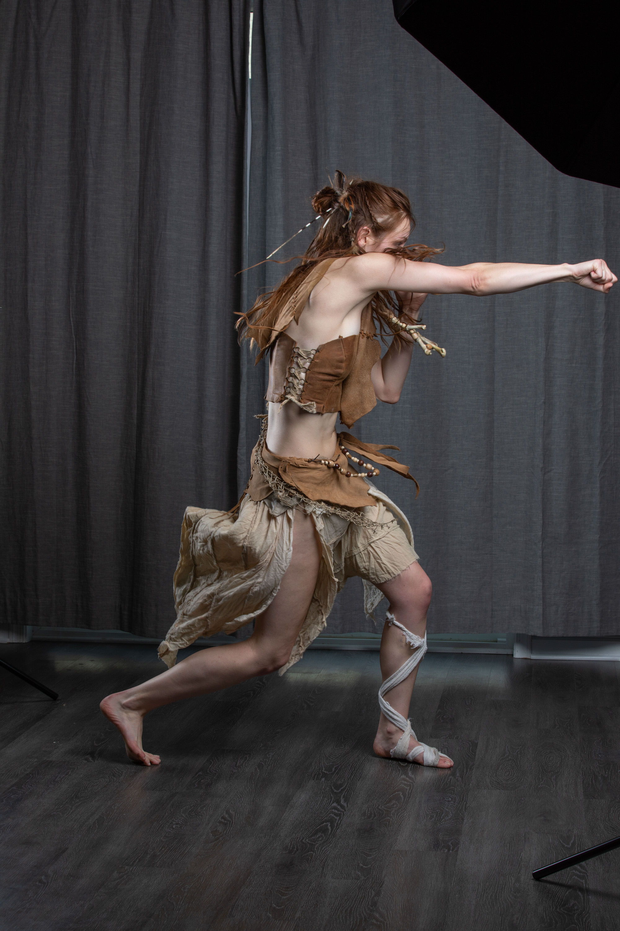 a woman in a tribalinspired costume strikes a dynamic fighting pose in a studio setting. she wears a brown leather top and skirt, her hair is styled with feathers, and her legs are wrapped in fabric. the background is a dark gray curtain, and the lighting is bright and focused on the subject. her powerful stance and determined expression convey a sense of strength and agility.
