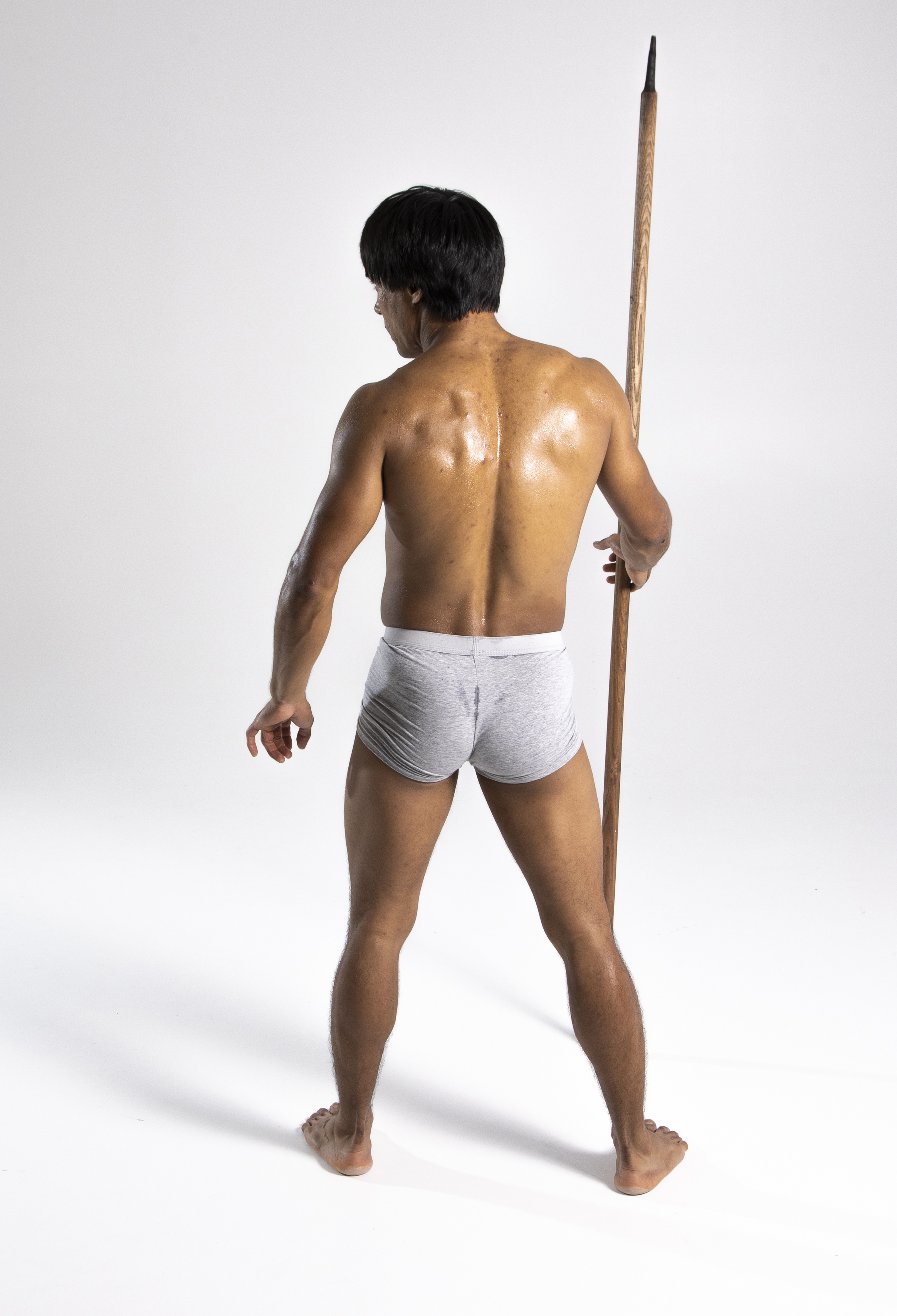 a muscular man stands with his back to the viewer, holding a spear. he is wearing grey underwear.