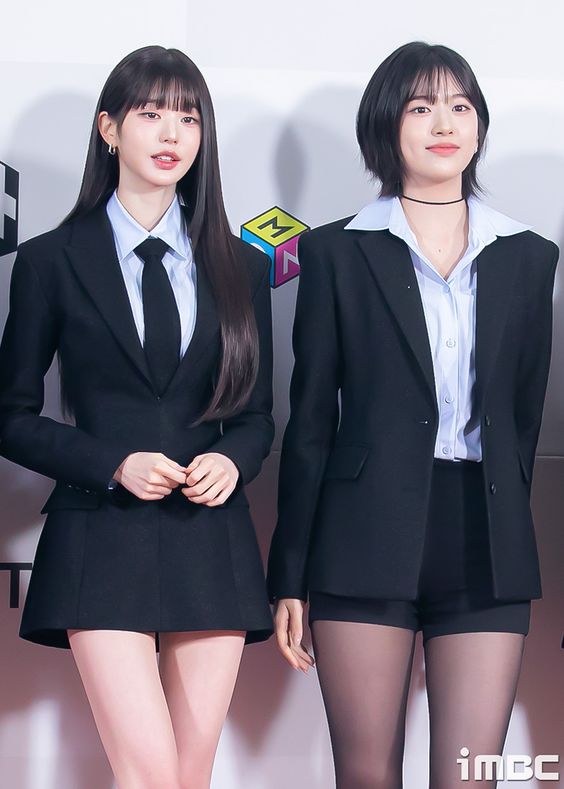 two young women are posing on a red carpet. they are both wearing black suits. the woman on the left has on a black tie and a white shirt. she is wearing a short skirt with her suit and has long black hair. the woman on the right is wearing a blue button down shirt under her jacket and a black tie. she is wearing black shorts with her suit and has short black hair. both women are smiling and looking at the camera.