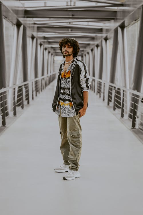 a full shot of a man standing in the center of a bridge, wearing a patterned shirt, a black and white jacket, and cargo pants.