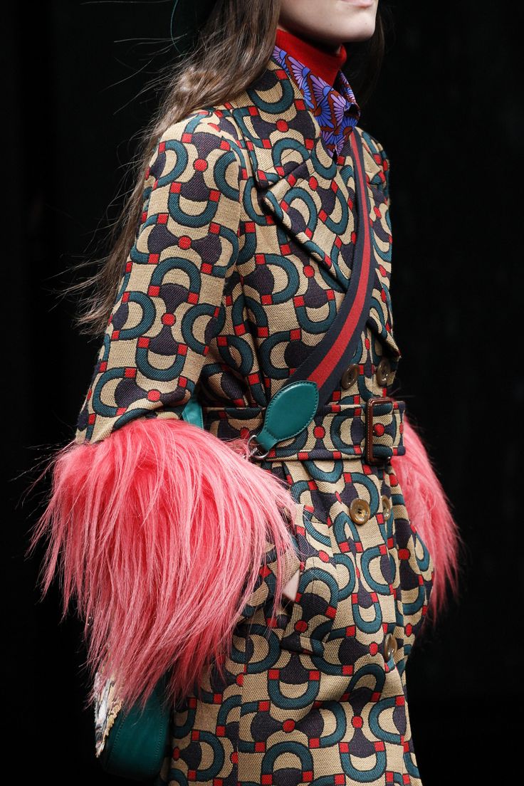 loseup detail shot of a model showcasing a gucci coat on a runway. the coat features a striking geometric pattern in teal, red, and beige, with pink fur cuffs.  a teal handbag with a red and black strap is partially visible. the model's hair is long and brown. the background is dark. the image focuses on the intricate details of the coat's design and texture.