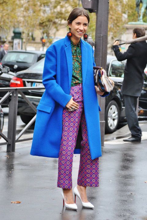 a stylish woman in a vibrant blue coat and patterned purple pants stands on a parisian street. she's wearing white high heels and large statement earrings, completing a fashionable autumn/winter look.  the photo captures her in a candid moment, showcasing street style elegance.