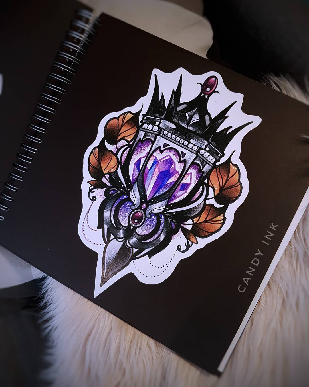 a black sketchbook lays open to a page with a sticker or drawing of a stylized  lantern in black, purple, and orange. the background of the page is black and in white it says "candy ink." the book is spiral bound on the left side and sits on a white, furry surface.