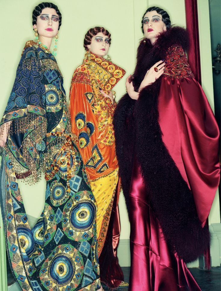 three women are standing close together wearing highly decorated, vibrant dresses that appear to be inspired by the 1920s fashion. the woman on the left has a long, flowing gown with intricate beadwork in shades of blue, green, and gold. the dress features a fringe detail along the hem. the woman in the middle wears an orange and yellow dress with detailed patterns and embellishments, her hair styled in tight finger waves. on the right, a woman sports a red satin dress trimmed with dark fur, and a beaded accent at the top, and also has a vintage hairstyle with dark makeup and red lipstick. all three women have similar, dramatic makeup with dark eyeliner and defined eyebrows. they exude a theatrical, vintage, and flamboyant style.