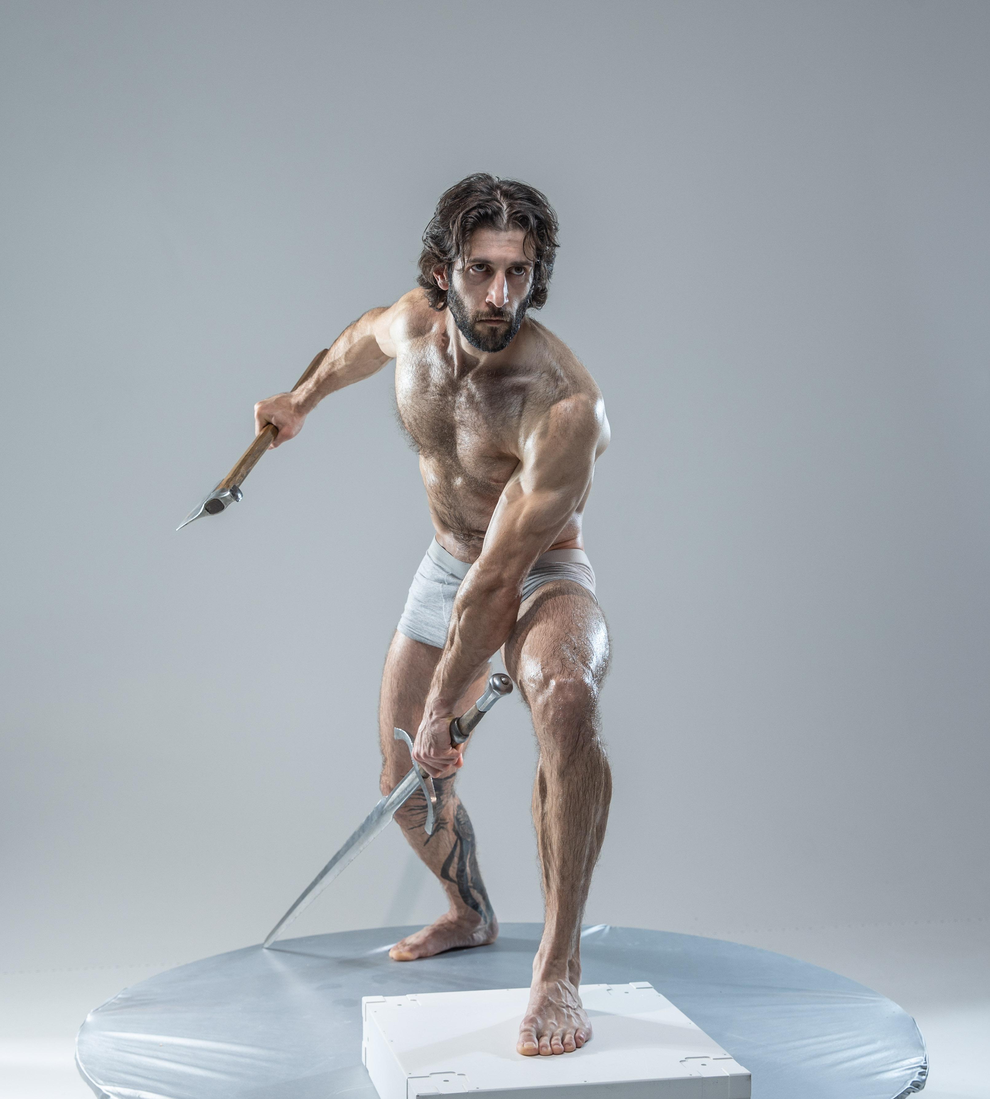 a muscular man, glistening with sweat, stands poised for battle in a studio setting. he holds a sword in his right hand, angled low, and an axe in his left, raised high. the image exudes power and action, capturing the intensity of the warrior's stance. he is barechested, wearing only white underwear, with his long hair flowing loosely. the background is a neutral gray, focusing all attention on the dynamic pose and physicality of the subject.