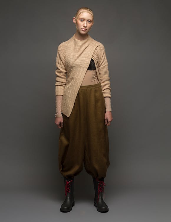 a fullbody studio shot features a female model against a neutral gray background, showcasing a unique fashion ensemble. the model wears a beige, asymmetrical sweater with textured knit details, paired with loosefitting, dark brown pants that balloon at the thighs. black boots with striking red laces complete the look. the model's expression is neutral, and her pose is centered, emphasizing the unconventional silhouette and textures of the clothing. the overall aesthetic suggests a blend of modern design with a touch of futuristic or avantgarde style.
