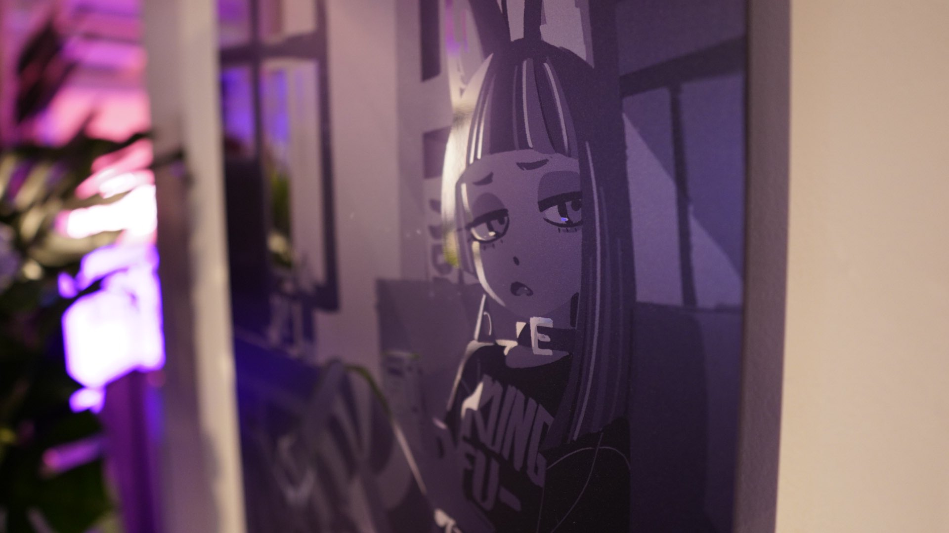 the image shows a framed illustration of an animestyle girl with bunny ears on her head. the girl has black hair with white streaks, dark eyes, and is wearing a black tshirt with white text that reads "hunte". she also has a black choker around her neck. the artwork is done in a black, white, and gray color scheme, with a purple hue cast on it from the surrounding environment. the image is taken from a slightly offcenter perspective, and the artwork appears to be hanging on a wall. there is a glimpse of a room in the background with purple lighting and some greenery visible on the left side of the image.