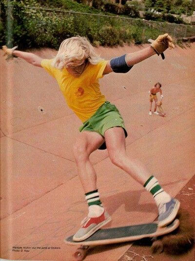 a young blonde girl in a yellow shirt and green shorts is skateboarding on a sunny day. she is wearing a pair of green and white socks and shoes. her hair is long and flowing in the wind. she is skateboarding down a street and she looks like she is having a lot of fun.