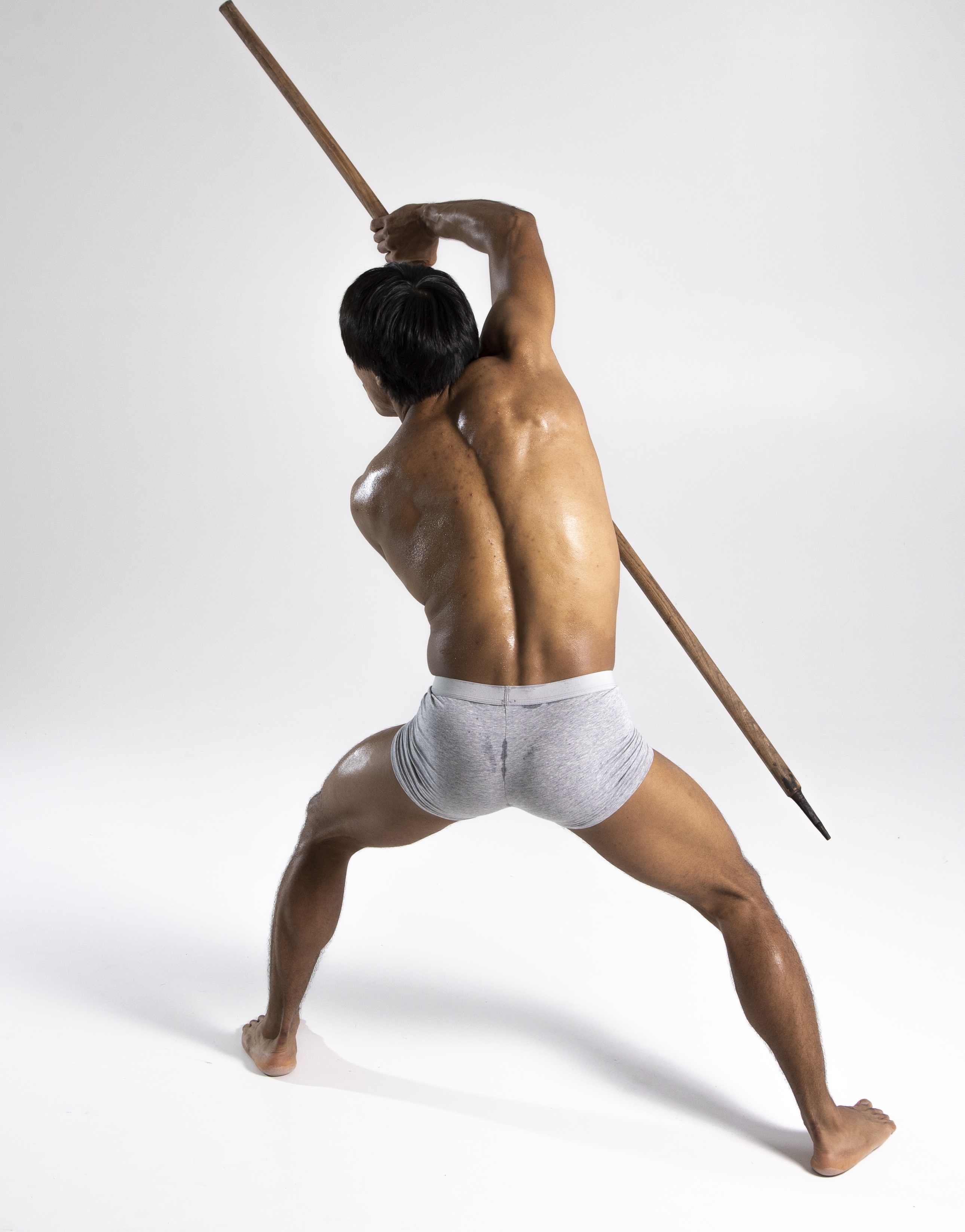 a shirtless man in shorts is holding a pole or spear, viewed from the back. he is muscular and in a stance, possibly training or exercising.