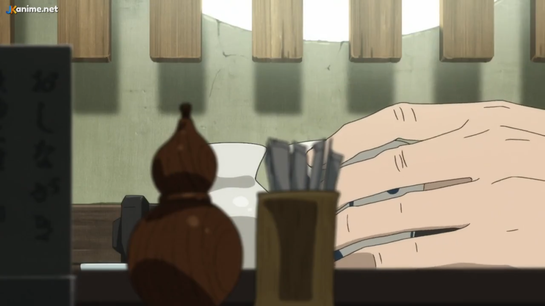 the image shows a closeup of a man's hand resting on a table. the hand is positioned near a small, white container filled with toothpicks. there's also a brown gourdshaped object on the table, and a wooden structure with vertical slats in the background.  the overall color palette is warm and muted.