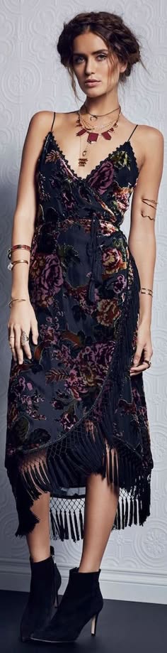 a woman with long brunette hair braided and a floral velvet dress is looking at the camera in a studio photoshoot. she is wearing a necklace with multiple chains and a pendant. there are multiple bracelets on her wrists. the dress has fringe detailing and she is wearing black boots.