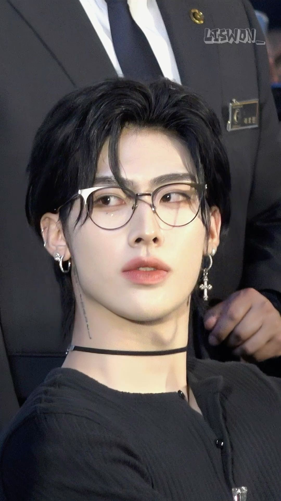 a  head and shoulders image of an asian male with black hair, wearing a black shirt and glasses with a silver chain. the background is blurred.