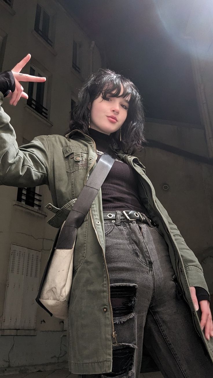 a woman stands outside and gives a peace sign. she is wearing a green jacket, black turtleneck shirt, black jeans with holes patched with black velvet, a black belt, and a shoulder bag. there are buildings in the background and she is wearing jewelry.