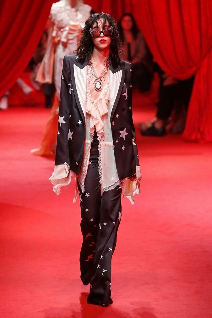 a woman in a black suit with white stars,  is walking on a red carpet runway. the suit is a black jacket over a white ruffle blouse.  the model is wearing black pants with white stars and black shoes.  she is also wearing silver jewelry, including a necklace with a large cameo pendant.  the woman has long dark hair and is wearing black sunglasses.  she is looking at the camera and posing for the photographer.  there are people in the background, partially blurred, but the focus is on the model.