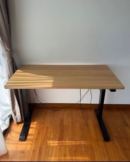 a wooden standing desk with black legs is set up in a room. the desk top is made of light brown wood with a slight grain pattern.  the room is made of hardwood floors. there is a light colored wall in the background and a curtain to the left.  the desk is set up with an electrical cord and a laptop.