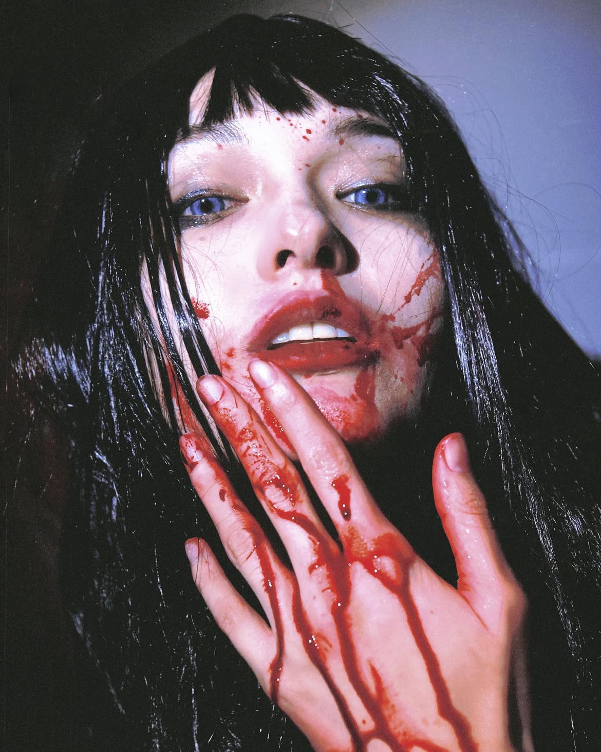 a closeup shot of a woman's face and hand covered in blood. she has dark hair with bangs, pale skin, and intense blue eyes. her lips are painted with red lipstick. the overall image evokes a dramatic, gory, and possibly horrorthemed atmosphere.