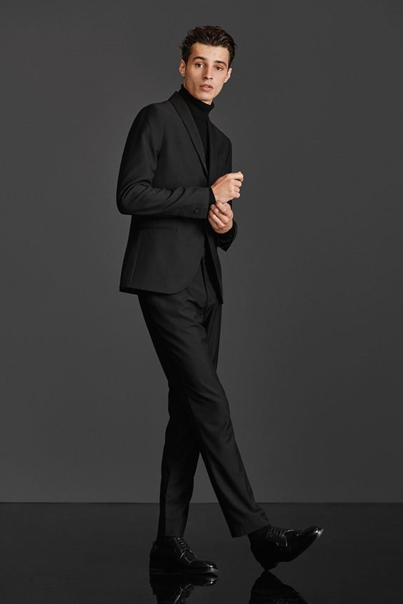 a male model is standing in a formal black suit with a black turtleneck. he has shiny black dress shoes on. the background is gray.