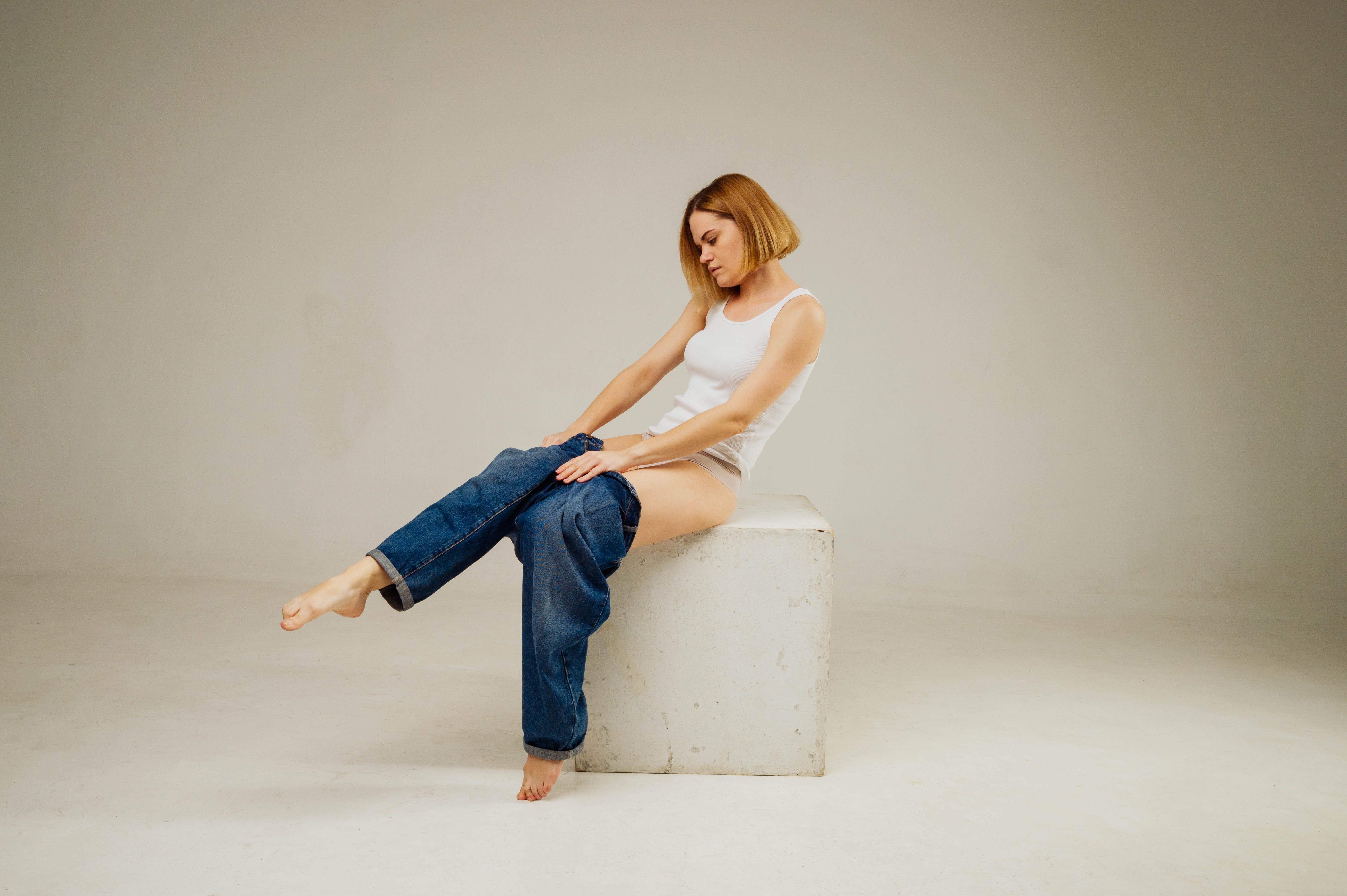a woman with short blonde hair, wearing a white tank top and underwear, is sitting on a white cube in a studio setting. she is in the process of putting on a pair of blue jeans. the image is welllit with soft, natural light. the overall feel is casual and minimal.