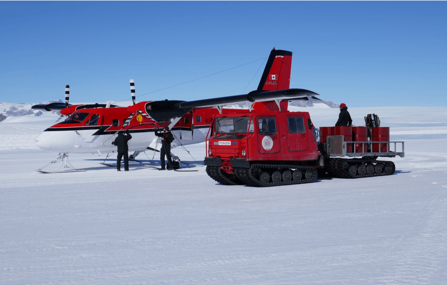 a red and white airplane sits on the snowcovered ground with a red truck and trailer behind it. two men in dark clothing are standing next to the airplane and a man is standing in the back of the truck. there are red barrels on the truck bed. the sky is a bright blue and mountains can be seen in the background. the scene looks like a scientific research expedition in antarctica.
