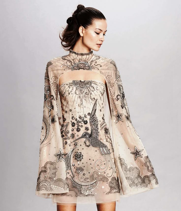 a woman is wearing a beige, short beaded dress with a capelike overlay featuring celestialthemed designs, including a hummingbird, stars, moon, and clouds. she has her hair in an updo. the dress has a high neckline and is embellished with delicate beads. the background is a soft neutral color, suggesting an indoor studio setting.