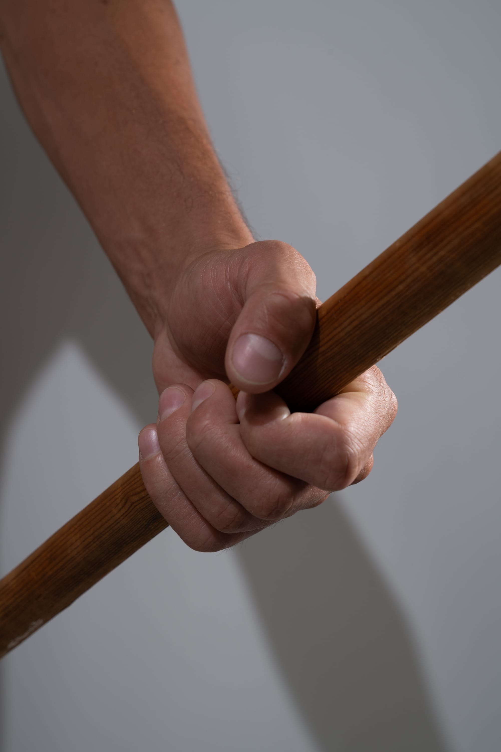 a closeup shot of a man's hand tightly gripping a wooden stick. the low angle perspective emphasizes the strength and tension in the hand and forearm. the neutral background and studio lighting keep the focus on the texture and details of the hand and wood.