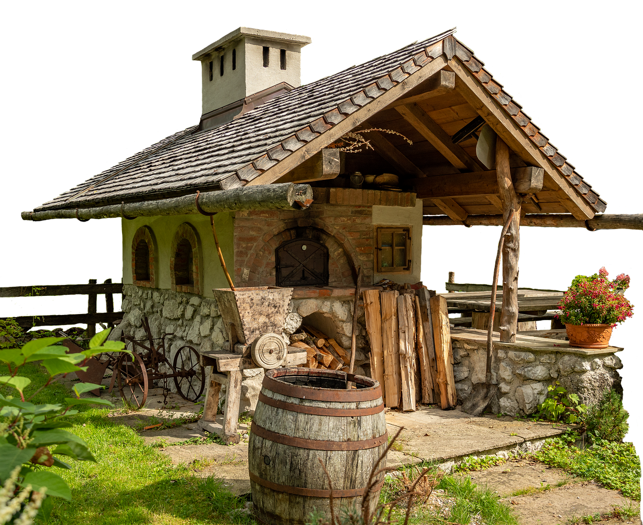an outdoor rustic oven with a chimney and covered shed is nestled in a garden. the oven is constructed of brick and stone, surrounded by wood and a barrel. flowers in a pot add color to the scene. the setting appears rural, with grass and a garden landscape.