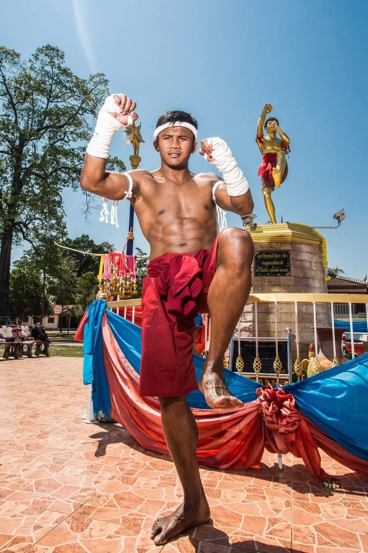 a full shot of a young, muscular muay thai fighter standing in a fighting stance in front of a statue. he is barefoot and wearing traditional red shorts and hand wraps. the background features a monument and some colorful fabric. the scene is bright and sunny, set outdoors on a paved surface. the fighter's physique and stance demonstrate his strength and readiness for combat. the statue in the background adds to the cultural context of the image.