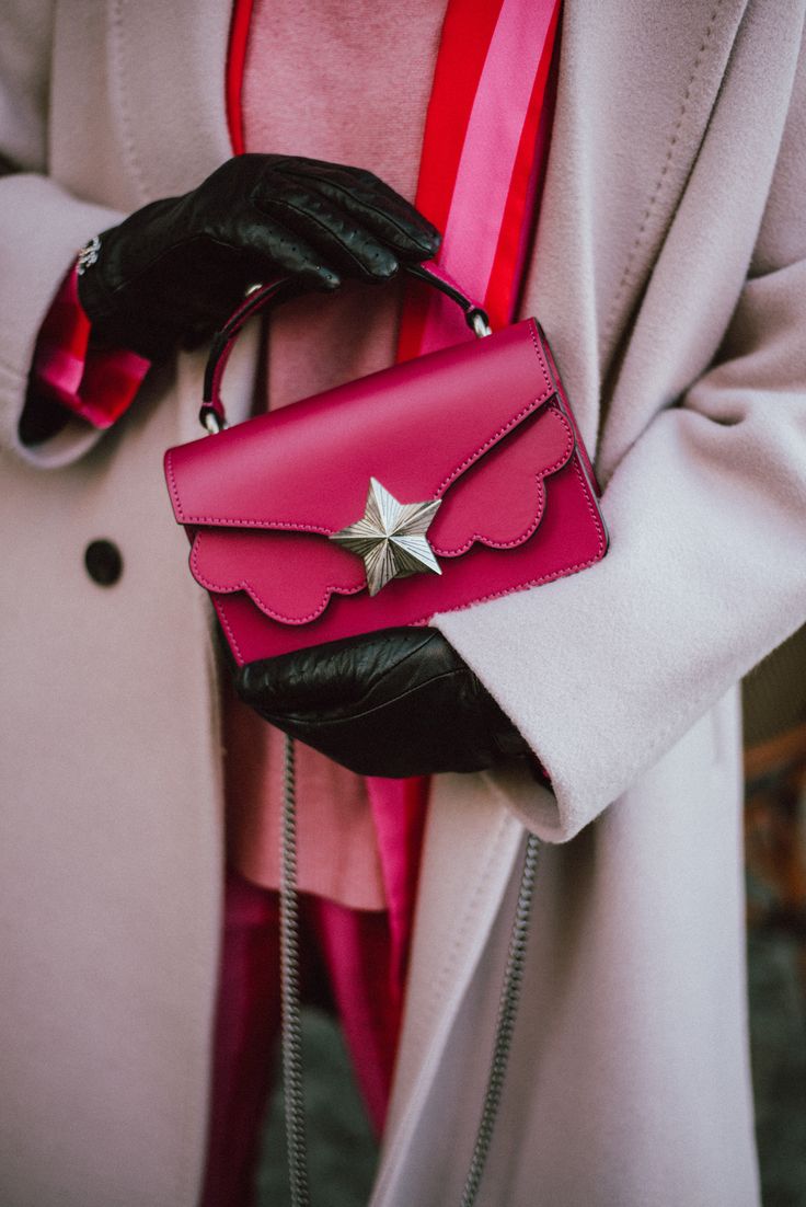 closeup shot of a woman wearing a light pink coat and black gloves, holding a vibrant pink handbag with a starshaped detail. the handbag has a unique scalloped edge and a chain strap. the woman is wearing a pink sweater underneath the coat.  the overall style is elegant and chic.