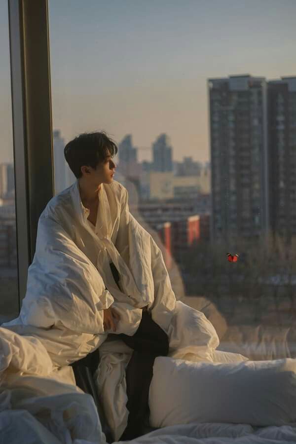 a young man sits on a bed facing a large window, gazing out at a city bathed in the warm light of a setting sun. he is wrapped in a white blanket, his expression contemplative. a single butterfly flutters in the distance, adding to the serene atmosphere. the room is minimally decorated, with white bedding and a neutral color palette that emphasizes the natural beauty of the cityscape outside.