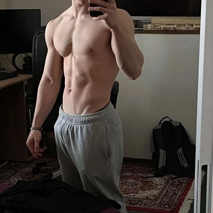 a muscular young man takes a mirror selfie in his room. he is shirtless, showing off his welldefined abs, and wearing grey sweatpants.  the background includes a desk with a computer, a chair, a backpack, and a rug on the floor. the overall lighting and style suggest a casual, possibly fitnessrelated, atmosphere.