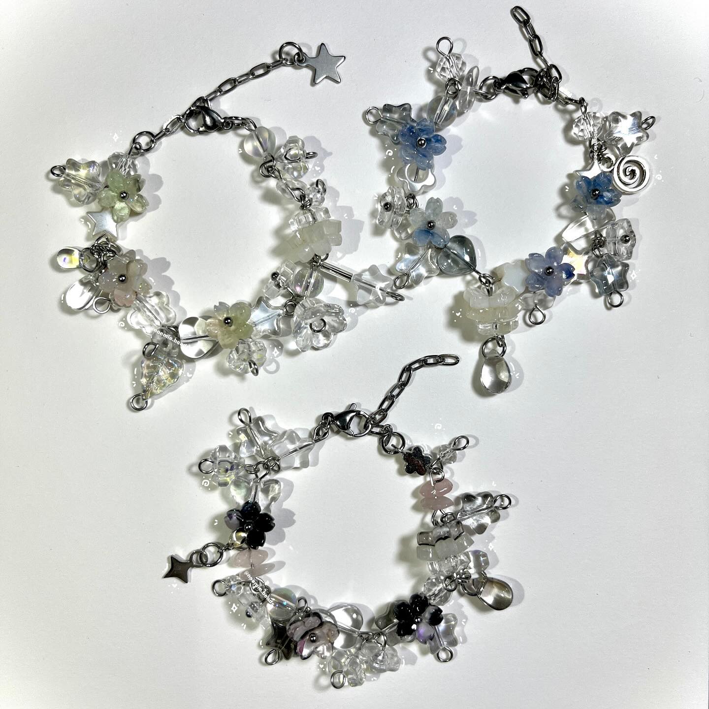 the image features three handmade beaded bracelets with floral and crystal bead accents. the bracelets are constructed with clear and iridescent crystal beads interspersed with flowershaped beads in various colors, including blue, green, pink, and purple. each bracelet has a metal chain closure with a clasp and a charm. one bracelet has a star charm, another has a water drop charm, and a third has a star charm. the bracelets are arranged on a white background.