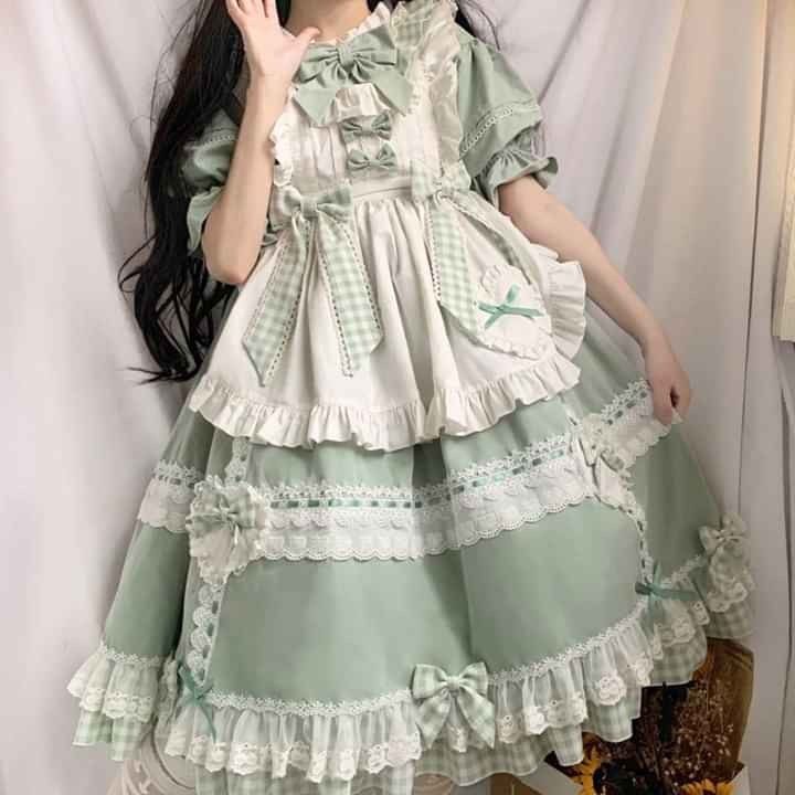 a young woman poses for a selfie while wearing a lolita fashion dress. the dress is a sage green color with white lace and gingham details. the dress has a tiered skirt and puffy sleeves, and is adorned with bows.