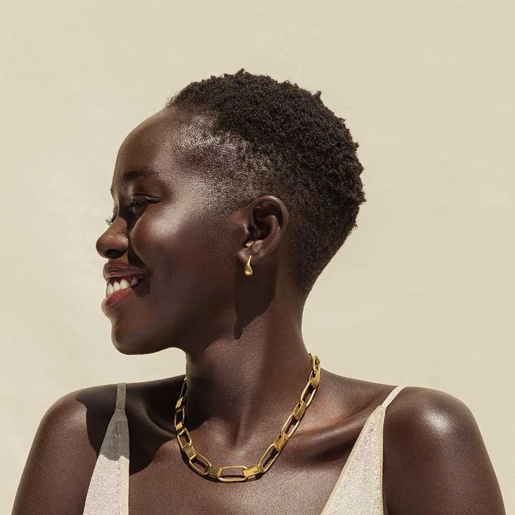 a woman with dark skin, short hair, and gold jewelry smiles in side profile against a neutral background. she is wearing a gold necklace and earrings.