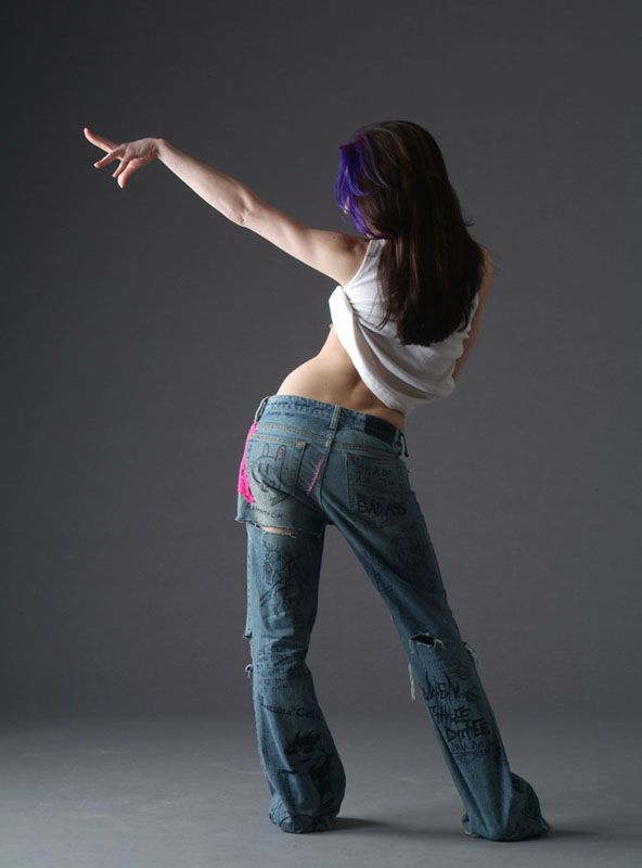 a woman with long, dark hair streaked with purple is captured middance move against a simple gray studio backdrop. she's wearing a white tank top that's riding up, revealing her toned midsection, and heavily distressed, lowrise jeans with black and pink designs. the soft lighting highlights her form and emphasizes the fluidity of her pose.