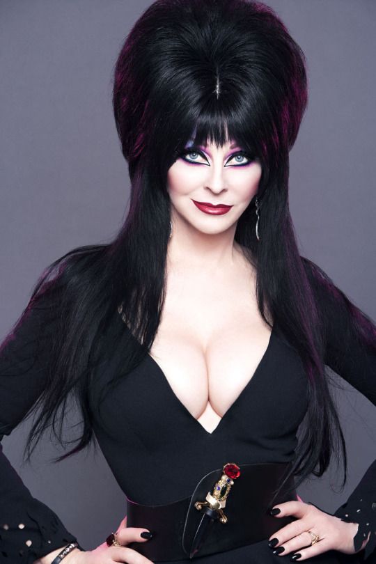 a portrait of a woman with long black hair styled in a beehive. she's wearing a black dress with a deep plunging neckline, a belt, and a small jeweled dagger pinned on her belt.  the woman is looking directly at the camera and has a serious expression.
