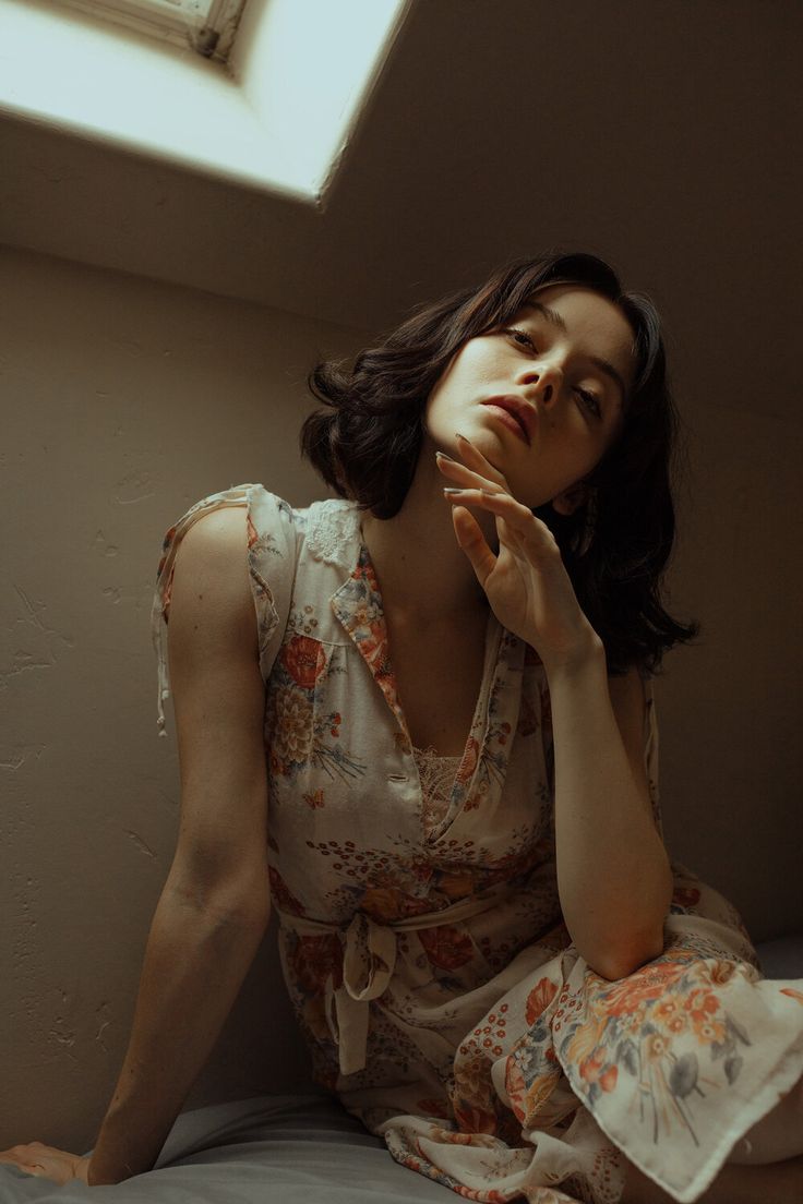 a woman in a vintage floral dress sits on a bed in a room, lit by natural light coming through a window. she looks thoughtfully toward the camera, her hand resting on her chin. the image has a soft, warm, nostalgic feel.