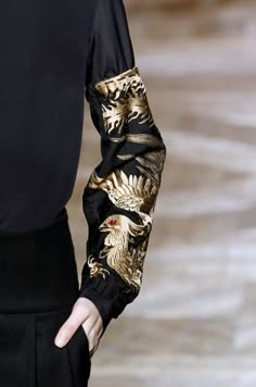closeup image of a black garment's embroidered sleeve showcasing a gold design with intricate details. the model's hand is visible holding the fabric, highlighting the elegant and luxurious quality of the clothing.