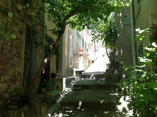 the image shows a narrow, sundappled alleyway in a mediterranean village. the alley is paved with cobblestones and features stone buildings on either side, some of which appear aged and rustic. lush greenery, including trees and various plants, overhangs the pathway, creating dappled shadows on the ground. the scene is peaceful and evokes a sense of quietude and charm typical of old european villages.  the alley appears to lead uphill, and the overall feel is one of a secluded and picturesque location.