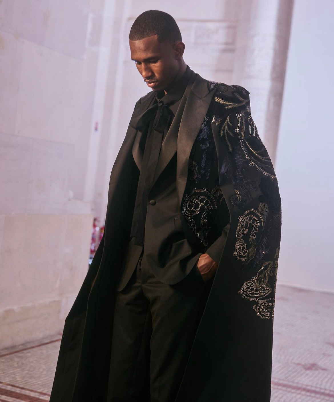 a man in a black suit with a black embroidered cape stands in a hallway with soft, natural lighting.  he is looking down, with his hand in his pocket. the image has an elegant and sophisticated mood.