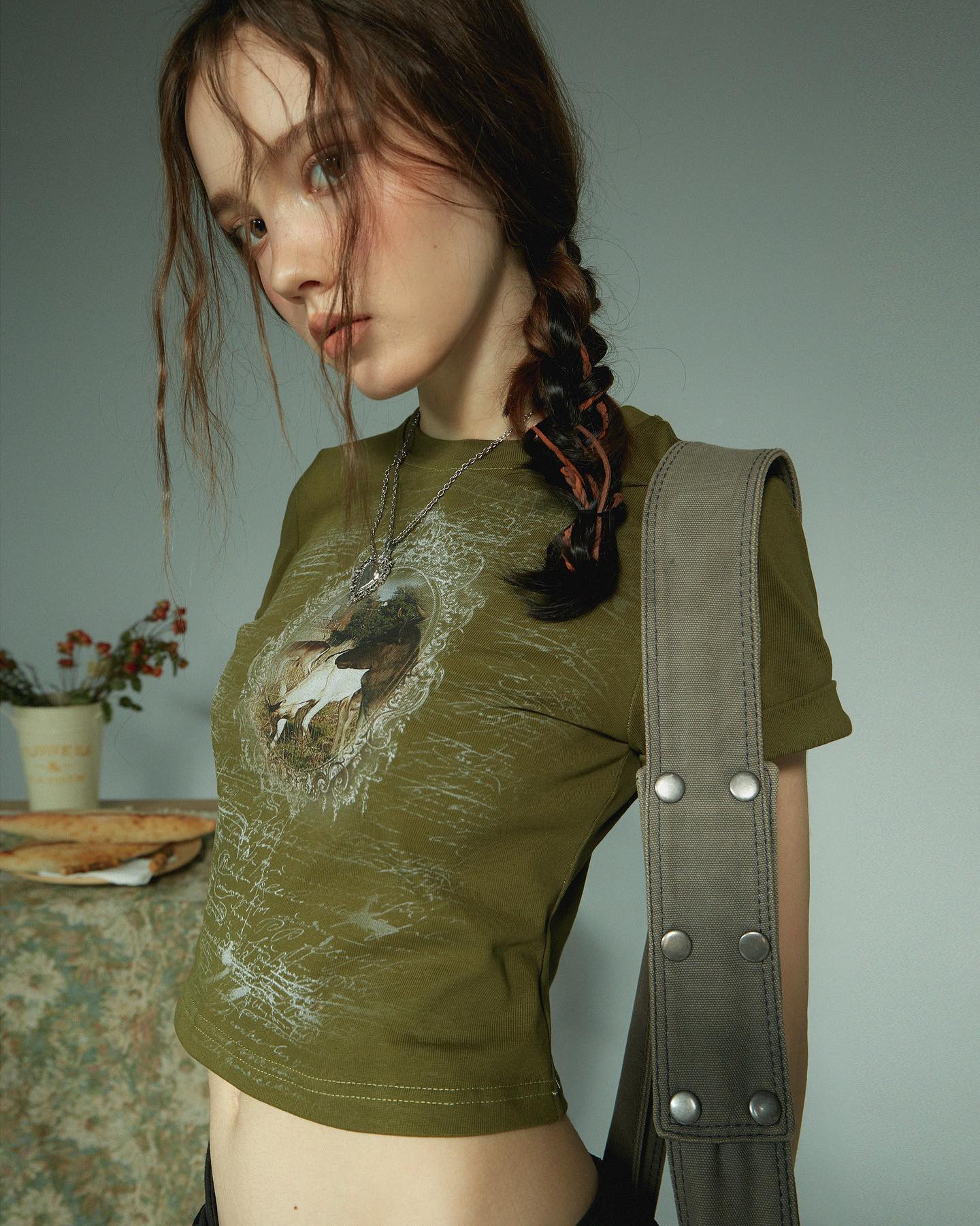 closeup shot of a young woman, possibly a model, wearing an olive green cropped tshirt with a vintagestyle graphic print. her long brown hair is styled in a braid.  she is also wearing a necklace and a grey canvas bag strap across her shoulder. the background is a muted grey, and the overall aesthetic is soft and somewhat nostalgic. the lighting is soft and even.