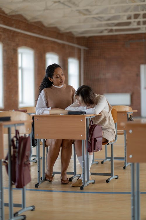 a young female student is sitting at a desk in a classroom, head down, appearing tired or upset.  an older female, possibly a teacher or mentor, is sitting beside her, offering comfort and support. they are sitting at wooden desks in a classroom with brick walls and large windows. backpacks are on the floor near the desks. the overall atmosphere suggests a supportive and caring learning environment.