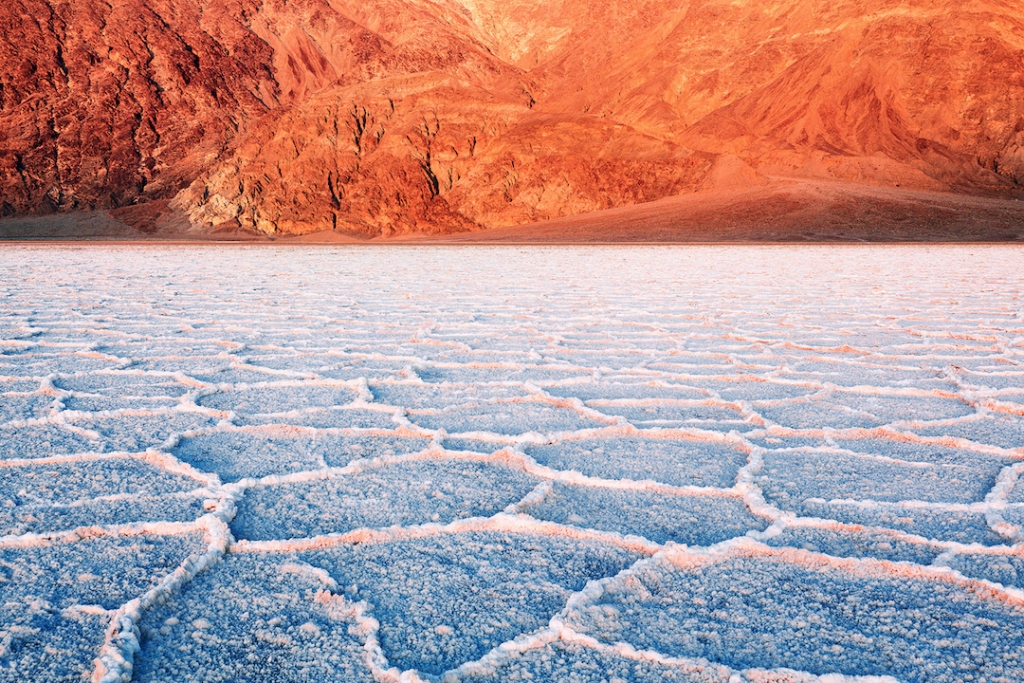 a scenic shot of death valley's salt flats, with the cracked earth of the badwater basin forming a mosaic pattern in the foreground. in the background, there is a reddishorange mountain range under a bright sky. the color palette of the image is a mix of blue, white, and warm earth tones, highlighting the contrast between the salt deposits and the surrounding geological formations.