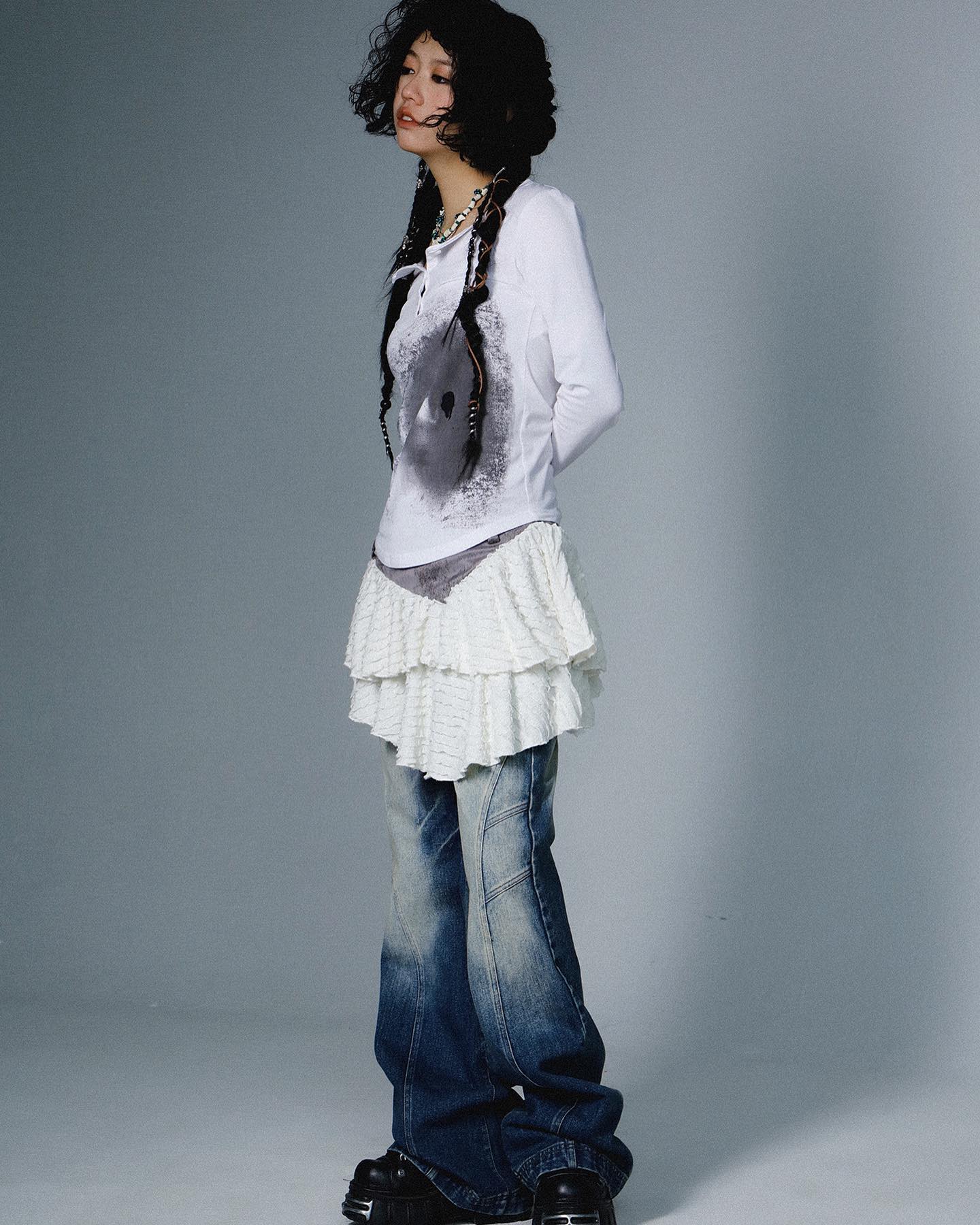 a fullbody studio shot of a young woman with dark, curly hair styled in braids. she is wearing a white longsleeved shirt with a dark graphic print, a white ruffled layered skirt, and distressed blue bellbottom jeans. she's also wearing chunky black boots and a simple necklace. the overall style is casual but with a grunge or alternative feel, potentially inspired by japanese fashion trends.  the background is a plain, light grey.  the woman is standing slightly angled away from the camera, with a thoughtful or pensive expression.