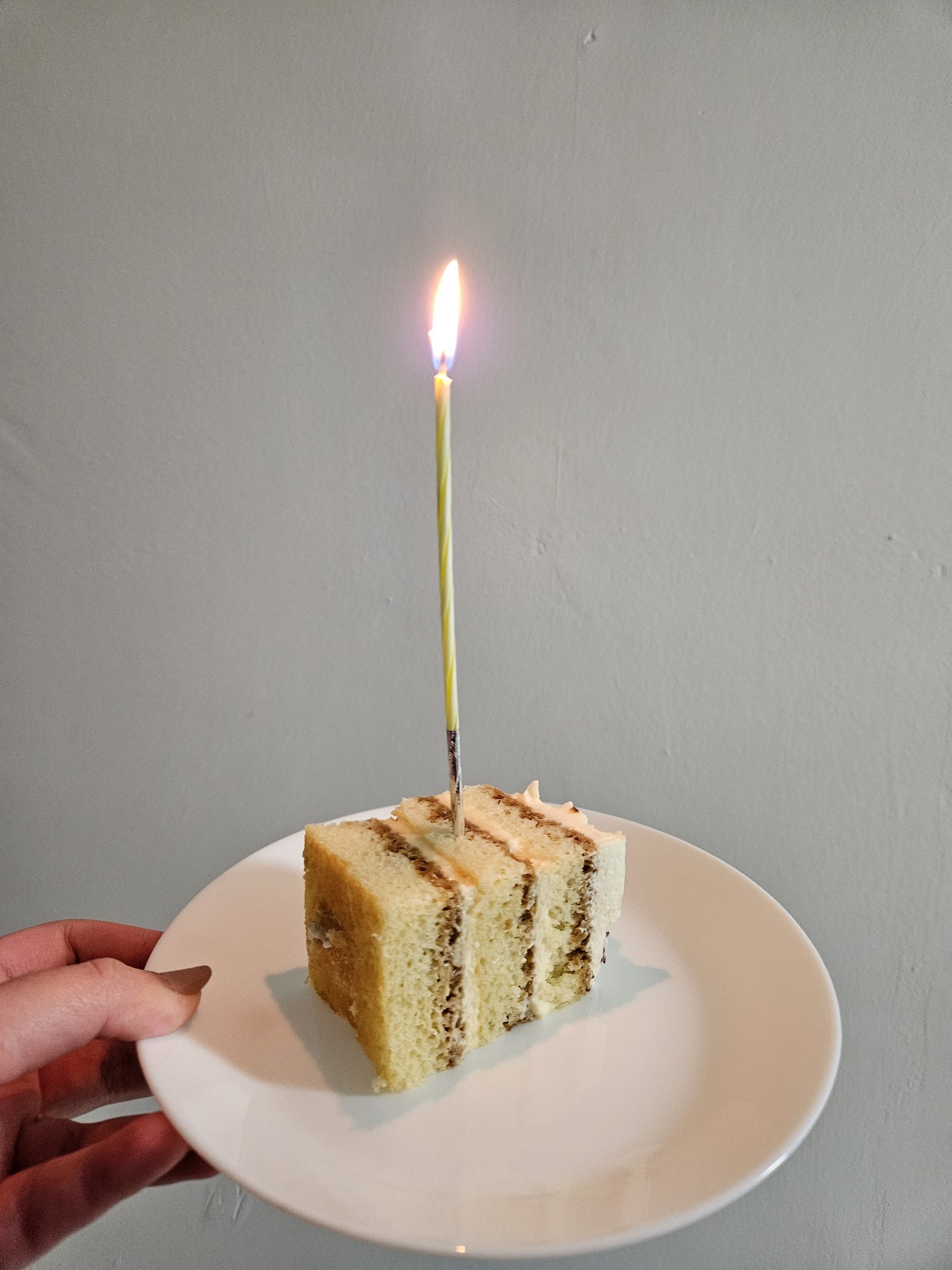 a slice of layered cake with a lit birthday candle on a white plate, held by a hand. the background is a plain, lightcolored wall.